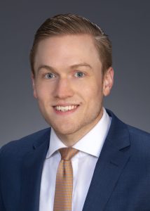 Professional headshot of a man wearing a blue blazer and gold tie, smiling against a neutral background.