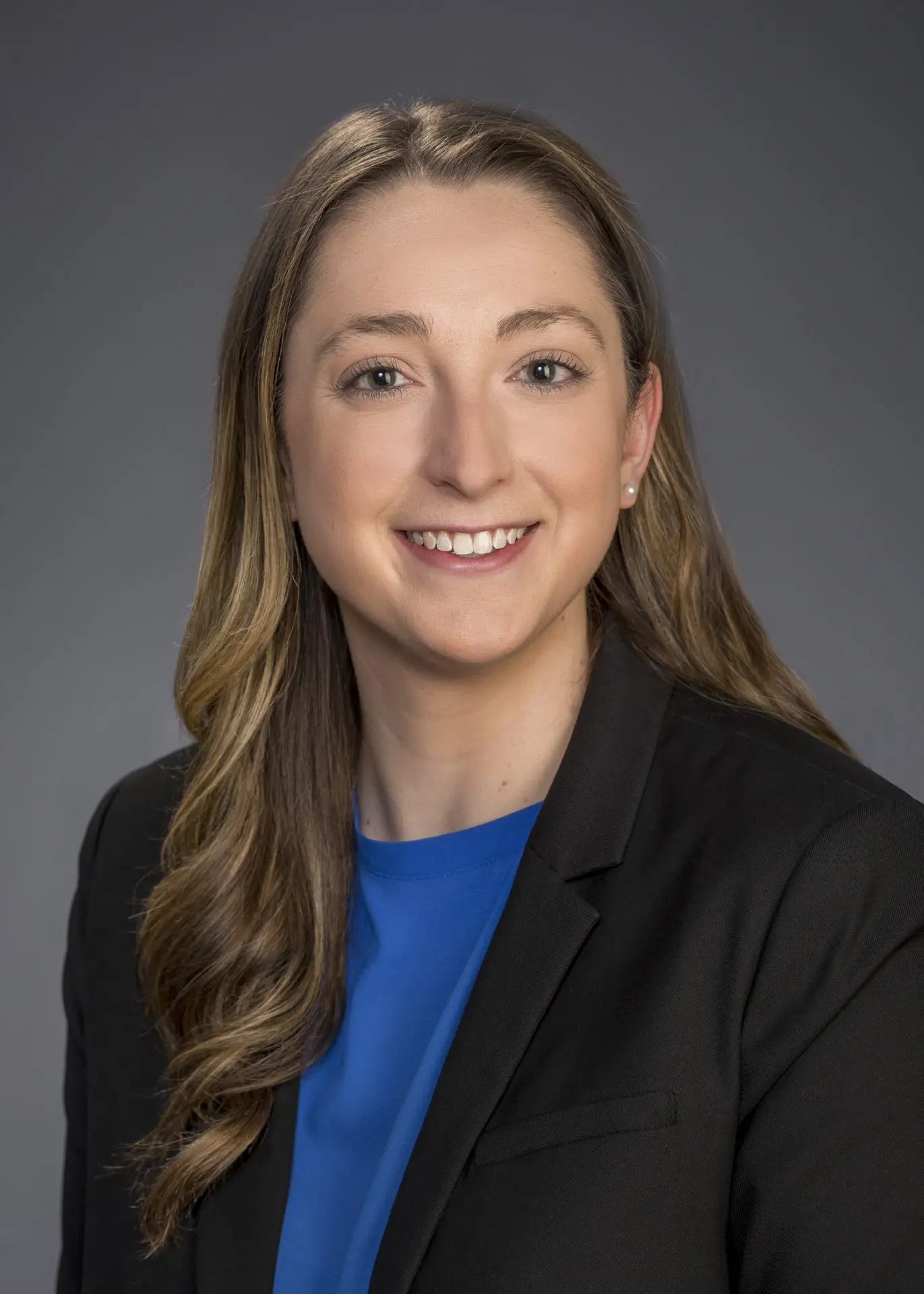 CJG_Graziadei_Christina_5x7 Professional headshot of Christina Graziadei with long wavy hair, wearing a black blazer and a blue blouse, smiling against a neutral gray background.