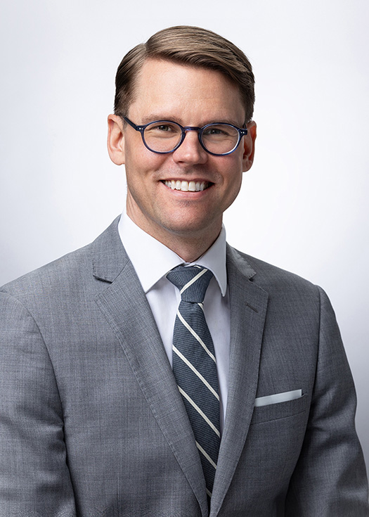 DanNorton_245x175 Smiling man in a gray suit, white shirt, striped tie, and blue-framed glasses against a light background.