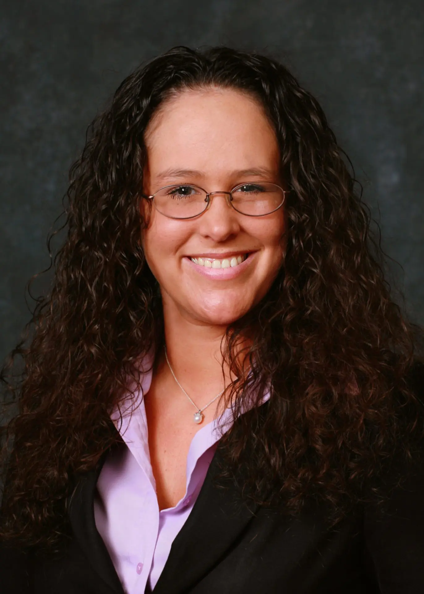 Professional headshot of a Erica Lawson with long curly hair, wearing glasses, a black blazer, and a lavender blouse, smiling against a dark studio backdrop.