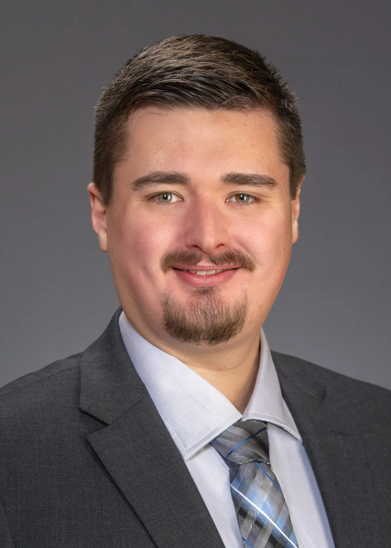 Professional headshot of a smiling man in a dark suit against a gray background.