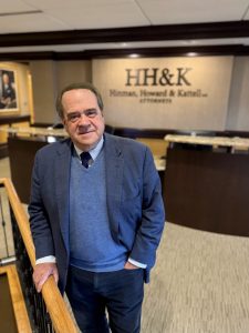 Man in a blue checkered blazer and sweater, standing on a staircase in a law firm lobby with an HH&K sign in the background.
